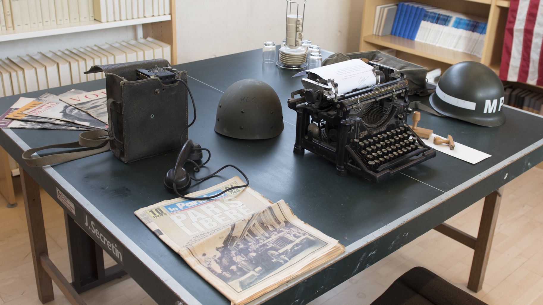Bureau imaginé au SHAPE, entre 1952 et 1967 : machine à écrire mécanique Underwood, casques militaires, vieux journaux.