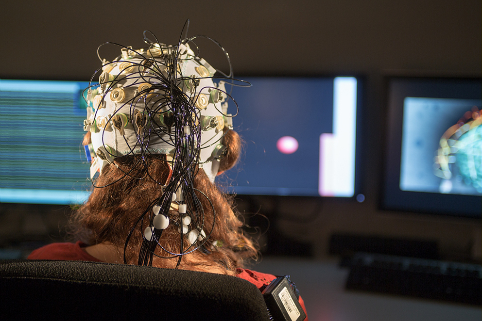 Photo d'une femme assise devant trois écrans d'ordinateurs, vue de dos. Elle porte un casque d’électroencéphalographie